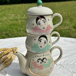 Puede incluir: Juego de té de cerámica apilada con acabado color crema. El juego incluye una tetera y dos tazas apiladas, cada una adornada con un diseño de cara de arlequín y detalles de estrellas. La tetera presenta el texto "Nous deux" y ribetes dorados.
