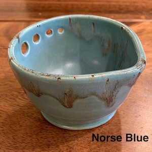May include: A ceramic bowl in a light blue glaze with brown drip accents along the rim and base. The bowl features three circular holes on one side of the rim and a small spout on the opposite side. The text Norse Blue is printed in black.