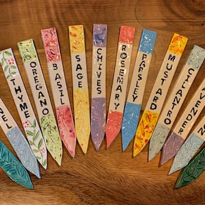 Puede incluir: Un conjunto de 14 marcadores de jardín de cerámica coloridos con diferentes nombres de hierbas y plantas escritos en ellos. Los marcadores están pintados con varios diseños florales y tienen un extremo puntiagudo para una fácil inserción en el suelo.