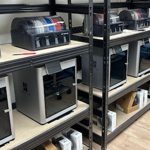Puede incluir: Una fila de estantes de metal negro con múltiples impresoras 3D y carretes de filamento. Las impresoras son plateadas y negras con una puerta de vidrio transparente. Los carretes de filamento son de varios colores, incluidos rojo, azul, morado y negro.