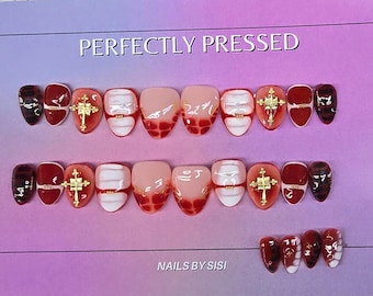 May include: A set of press-on nails featuring a mix of red, white, and nude tones with a grid pattern, gold cross charms, and a glossy finish. The text PERFECTLY PRESSED and NAILS BY SISI is visible on the purple background behind the nail set.