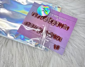 May include: A set of press-on nails featuring a deep maroon base with a white, web-like pattern. The nails are displayed on a purple card labeled "PERFECTLY PRESSED" and "NAILS BY SIS," topped with a holographic "Thank you" sticker inside a shiny pouch.