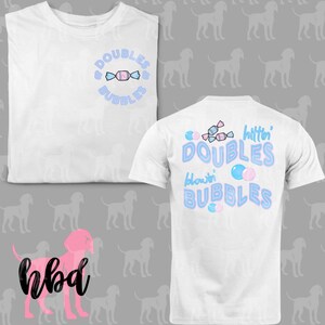Puede incluir: Camiseta blanca con gráficos de caramelos azules y rosas y burbujas. La parte delantera de la camiseta dice "Double & Bubbles". La parte trasera de la camiseta dice "Bitin' Doubles Blowin' Bubbles".