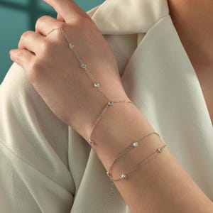 May include: A silver-toned hand chain featuring a delicate metal link chain adorned with small, round, clear stones. The jewelry piece connects a bracelet section around the wrist to a ring component that loops around the middle finger.