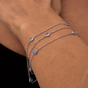 May include: A silver bracelet with three delicate chains, each adorned with round, light blue gemstones. The bracelet is shown on a wrist, with the gemstones catching the light. The jewelry is simple and elegant.