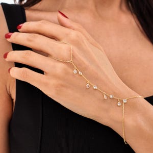 Peut inclure: Une chaîne de main dorée composée d'une fine chaîne reliant une bague à un bracelet de poignet. La chaîne est ornée de plusieurs petites pierres transparentes de taille ronde qui pendent à intervalles réguliers le long de la main.