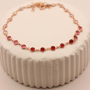 May include: A delicate gold-toned bracelet featuring a series of alternating pink and red gemstones. The bracelet is displayed on a white, textured surface. The gemstones are round and evenly spaced along the chain.