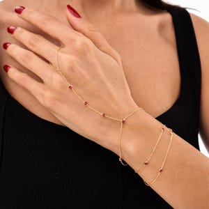 Op de afbeelding: Een gouden handkettingarmband met rode edelstenen. Het delicate sieraad verbindt met een ring en wikkelt zich om de pols met een dubbele ketting, met meerdere rode stenen.