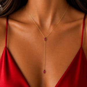 May include: A gold lariat necklace featuring two marquise-cut red gemstones. The chain forms a Y-shape, with one stone positioned at the centre junction and a second stone hanging at the end of the vertical drop, creating a delicate layered look.