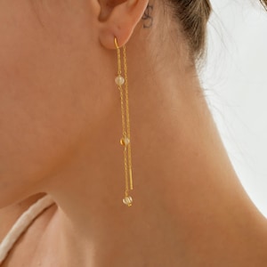 Könnte beinhalten: Goldfarbene Kettenohrringe mit drei kleinen, runden, hellfarbigen Perlen und einem kleinen Stab. Die Ohrringe sind lang und hängen herab, mit einem filigranen Design. Die Ohrringe werden am Ohr einer Person gezeigt.