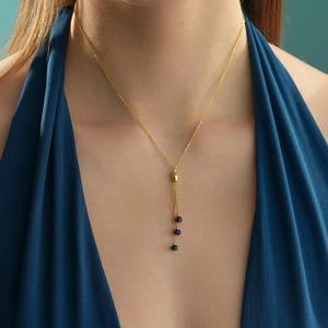 May include: A gold-tone chain necklace featuring a central vertical drop element. The drop consists of a small gold bead followed by three dangling, multi-coloured spherical beads in shades of deep red, dark blue, and green, suspended on thin gold chains.