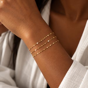 May include: A delicate gold chain bracelet featuring a series of small, flat, reflective metallic links. The bracelet is designed with a repeating pattern of tiny, shimmering segments that catch the light, creating a continuous, elegant, and refined appearance.