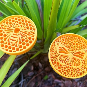 May include: Two round, yellow decorative coasters. One features a bee design, the other a butterfly. Both have a honeycomb pattern background. The coasters are set against a backdrop of green leaves.