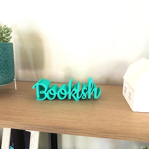 May include: A teal 'Bookish' word art ornament is displayed on a wooden shelf. A teal patterned plant pot with a succulent and a white house ornament are also on the shelf. Ideal for a home library or book lover's space.