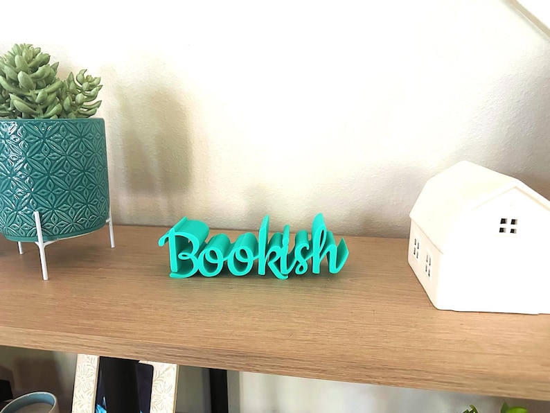 May include: A teal 'Bookish' word art ornament is displayed on a wooden shelf. A teal patterned plant pot with a succulent and a white house ornament are also on the shelf. Ideal for a home library or book lover's space.
