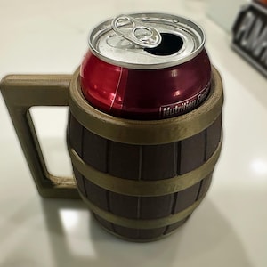 May include: A brown barrel-shaped can holder with a handle, holding a red soda can. The holder has gold-colored bands and a handle. The can is open, showing the silver pull tab. The holder is on a white surface.