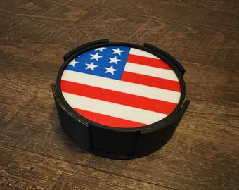 May include: A round coaster featuring the American flag design with red and white stripes and blue field with white stars. The coaster sits in a black holder. The coaster is a patriotic home decor item.
