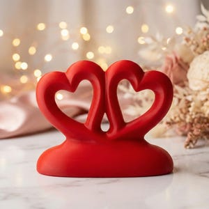 May include: A vibrant red sculpture featuring two intertwined hearts, symbolizing love and connection. The smooth, glossy finish of the hearts contrasts with the soft, blurred background of fairy lights and flowers. A romantic and decorative item.