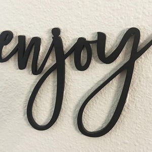 May include: Black cursive wooden sign spelling the word "enjoy". The sign is mounted on a white textured wall. The word is in a flowing script font.