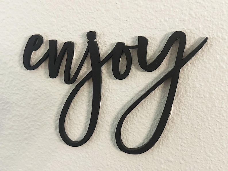 May include: Black cursive wooden sign spelling the word "enjoy". The sign is mounted on a white textured wall. The word is in a flowing script font.