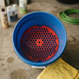 May include: A blue bucket with a red honeycomb-patterned insert, filled with water. A yellow microfiber towel rests beside the bucket. Cleaning supplies and a green hose are visible in the background. The bucket is likely for car washing or detailing.