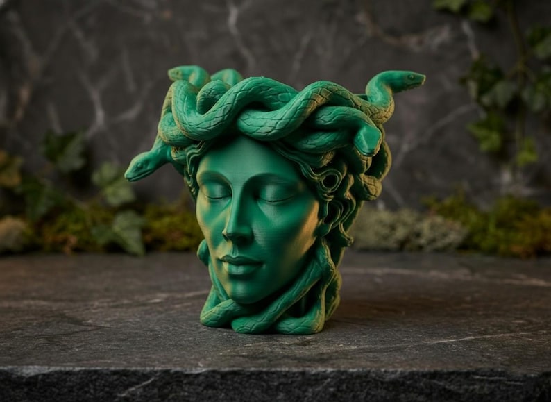 K&ouml;nnte beinhalten: Eine gr&uuml;ne Medusa-Kopf-Skulptur mit geschlossenen Augen und Schlangen als Haare. Die Skulptur besteht aus einem festen Material und steht auf einer dunkelgrauen Steinoberfl&auml;che. Der Hintergrund ist dunkelgrau mit gr&uuml;nem Laub.