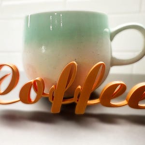 May include: A ceramic coffee mug with a gradient of colours, from light green to white, with an orange cursive "coffee" sign in front. The mug has a large handle and sits on a dark surface.