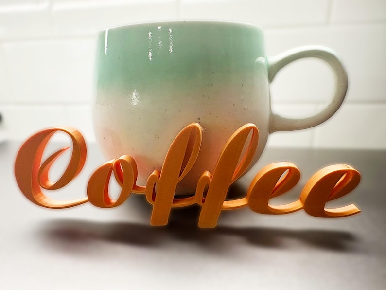 May include: A ceramic coffee mug with a gradient of colours, from light green to white, with an orange cursive "coffee" sign in front. The mug has a large handle and sits on a dark surface.