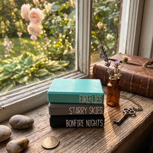 May include: A stack of three rectangular wooden blocks with smooth edges. The top block is teal with the text FIREFLIES, the middle block is gray with STARRY SKIES, and the bottom block is black with BONFIRE NIGHTS printed in white.