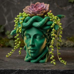 May include: A green Medusa head planter with a succulent and trailing plants. The planter features a detailed face with closed eyes and snakes for hair. The succulent is a light purple color, and the trailing plants are green.