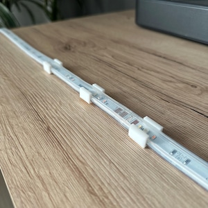 Puede incluir: Tira de luz LED blanca fijada a una superficie de madera con clips de plástico transparente. La tira se extiende a lo largo de la superficie.