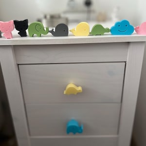 Può includere: Comodino bianco con tre cassetti. Ogni cassetto ha una manopola a forma di animale diversa. Il cassetto superiore ha una manopola di tartaruga gialla, il cassetto centrale ha una manopola di dinosauro gialla e il cassetto inferiore ha una manopola di elefante blu.