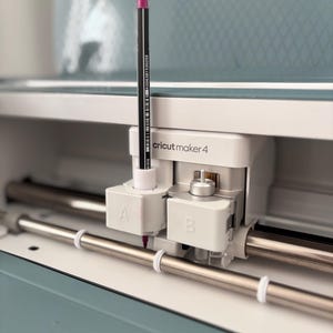 Op de afbeelding: Een close-up van een Cricut Maker 4 machine met een zwarte en roze pen erin. De machine is wit en lichtblauw, met zilveren metalen staven. De pen zit in de 'A'-sleuf.