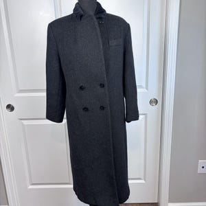 May include: A long, dark charcoal gray wool-blend overcoat featuring a double-breasted front closure with four visible black buttons, a high collar, long sleeves, and a single welt pocket on the upper left chest area, displayed on a mannequin.