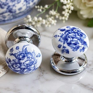 Könnte beinhalten: Zwei weiße Türknöpfe aus Keramik mit blauem Blumenmuster. Die Knöpfe haben einen polierten Chromsockel und werden auf einer Marmoroberfläche präsentiert. Das Blumenmuster zeigt detaillierte blaue Blumen und Blätter.