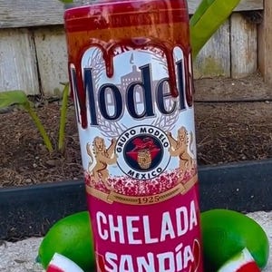 May include: A red and white insulated tumbler with a Modelo beer can design. The tumbler has a red lid and a straw. The tumbler is decorated with watermelon slices and lime wedges. The text on the tumbler reads "Chelada Sandia Picante".