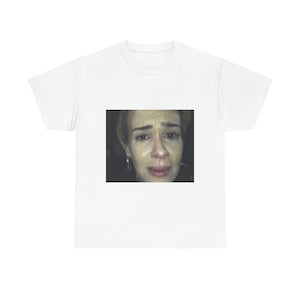 Może przedstawiać: Biały t-shirt z nadrukowanym obrazem kobiety o smutnym wyrazie twarzy. Kobieta ma blond włosy i nosi czarną koszulę.