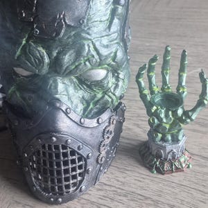 May include: A textured green mask with a dark metallic lower face covering featuring a grid pattern and gear accents, positioned next to a small green skeletal hand sculpture mounted on a stone-like base with red vine details.