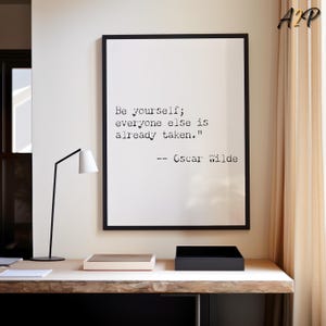 Può includere: Una stampa incorniciata con la citazione "Sii te stesso; tutti gli altri sono già presi" di Oscar Wilde. La stampa con cornice nera è appesa sopra una scrivania di legno con una lampada nera e due scatole.