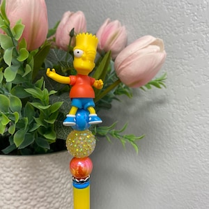 May include: A yellow pen with a Bart Simpson figure on top. The pen features a yellow barrel, a series of decorative beads in yellow and orange, and a white tip. The Bart Simpson figure is wearing a red shirt and blue shorts.