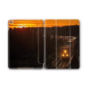 Könnte beinhalten: Eine iPad-Hülle mit einem Sonnenuntergang über einem Wald und einem Zug auf Gleisen. Die Scheinwerfer des Zuges leuchten, und der Himmel ist leuchtend orange. Die Hülle dient zum Schutz des iPads.