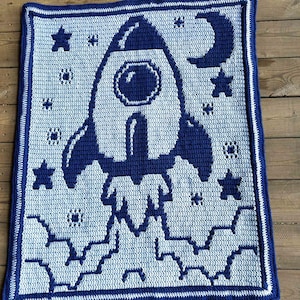 May include: A crocheted blanket featuring a blue and white rocket ship design, surrounded by stars and a crescent moon. The blanket has a blue border and is made of soft yarn. The overall theme is space and celestial bodies.