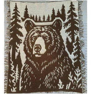 Könnte beinhalten: Eine gehäkelte Decke in Braun und Creme mit einem Bärenmuster. Der Bär steht im Mittelpunkt, umgeben von Bäumen. Die Decke hat einen Fransenrand und wird auf einer gestreiften Oberfläche präsentiert.