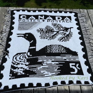 May include: A black and white crochet blanket featuring a design of a Canadian postage stamp. The stamp design includes the words "CANADA POSTES + POSTAGE", a loon, and the number "5¢". The blanket has a fringe border.