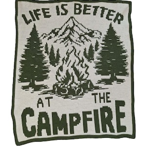 May include: A crocheted blanket with a forest scene and the text "LIFE IS BETTER AT THE CAMPFIRE." The design features a mountain, trees, and a campfire in shades of green on a cream background. The blanket has a green border.