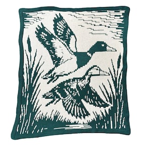 May include: A square, crocheted blanket featuring a teal and white design of two ducks in flight over a marsh. The blanket has a teal border and depicts reeds and water in the background. The ducks are detailed with white and teal stitches.