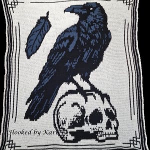 May include: A crocheted blanket featuring a raven perched on a skull, with a feather detail. The design is in shades of black and blue on a white background, framed by a black border. The text "Hooked by Kari" is visible at the bottom.