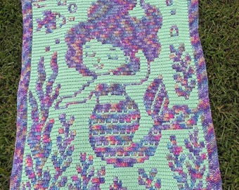 May include: Crocheted blanket featuring a mermaid design. The blanket has a light green background with a mermaid in shades of purple, pink, and blue. The border and details, including seaweed and bubbles, are also in shades of purple and pink.
