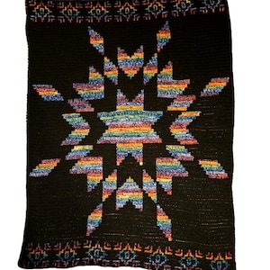 May include: A black crocheted blanket with a large, colorful starburst design in rainbow hues. The blanket has a geometric border with repeating patterns in orange, blue, and green. The blanket is made of soft yarn.