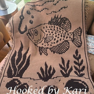 May include: A tan and brown crocheted blanket featuring a large fish design with a fishing hook, bubbles, and aquatic plants. The blanket has a fringed edge and the text "Hooked by Kari" is displayed at the bottom.